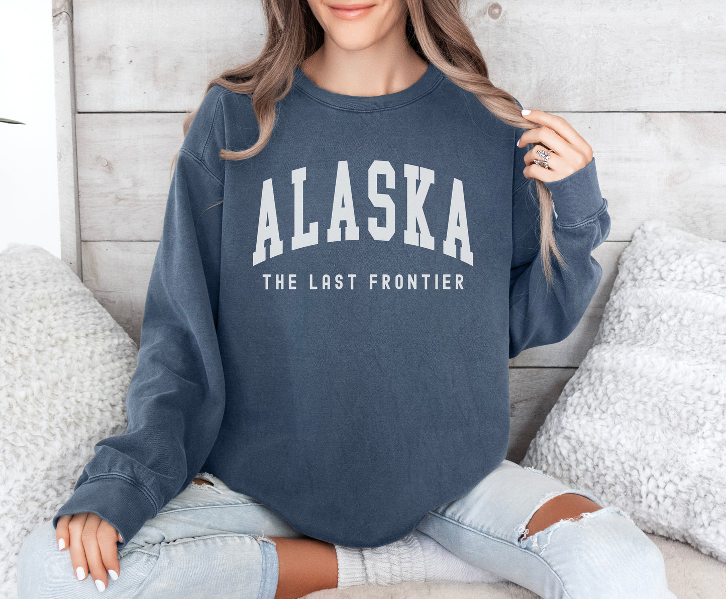 a woman sitting on a bed wearing a sweatshirt that says alaska the last frontier